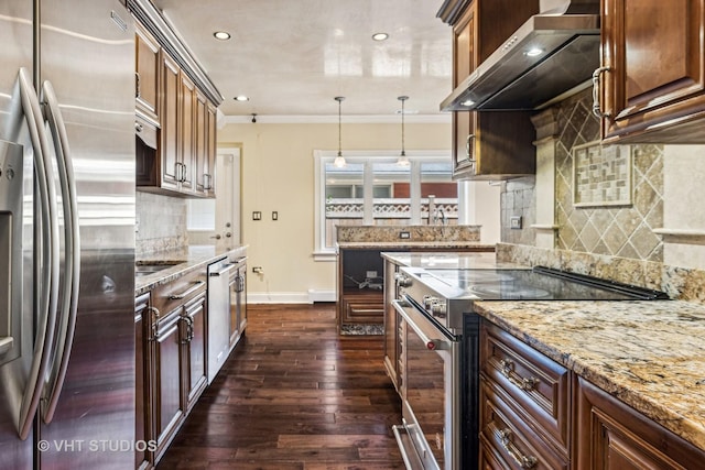 kitchen with dark wood-type flooring, appliances with stainless steel finishes, hanging light fixtures, light stone counters, and wall chimney exhaust hood