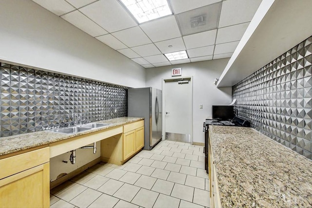 kitchen with a drop ceiling, freestanding refrigerator, light brown cabinetry, black electric range, and a sink