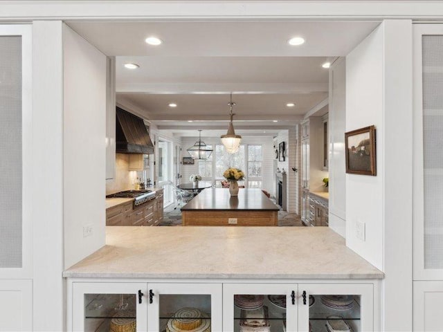 kitchen with a center island, wall chimney exhaust hood, beamed ceiling, glass insert cabinets, and stainless steel gas stovetop