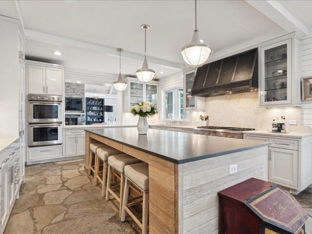 kitchen with decorative backsplash, double oven, stone tile flooring, premium range hood, and a sink