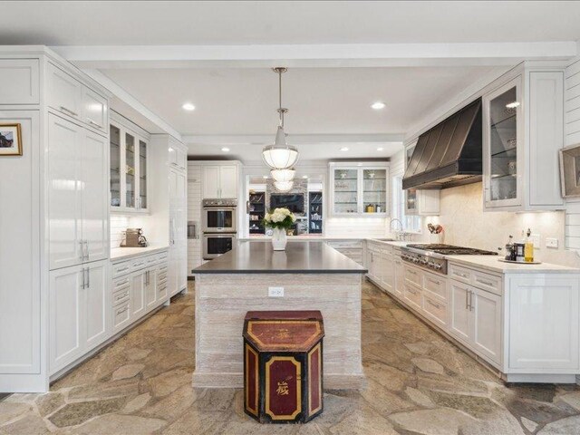 kitchen featuring stone tile floors, custom range hood, backsplash, appliances with stainless steel finishes, and white cabinetry