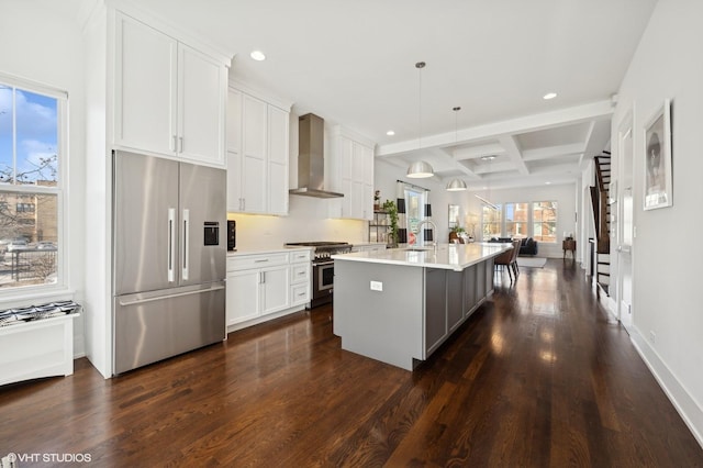 kitchen featuring wall chimney exhaust hood, coffered ceiling, high end appliances, decorative light fixtures, and a kitchen island with sink