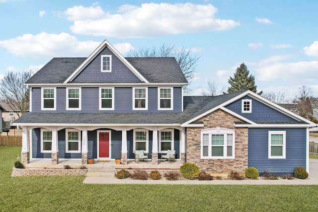 craftsman house with covered porch, fence, stone siding, roof with shingles, and a front yard