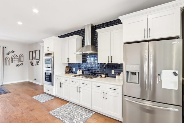 kitchen featuring light wood-style flooring, stainless steel appliances, light countertops, wall chimney exhaust hood, and tasteful backsplash