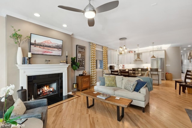 living room with crown molding, a fireplace, ceiling fan with notable chandelier, and light wood-type flooring