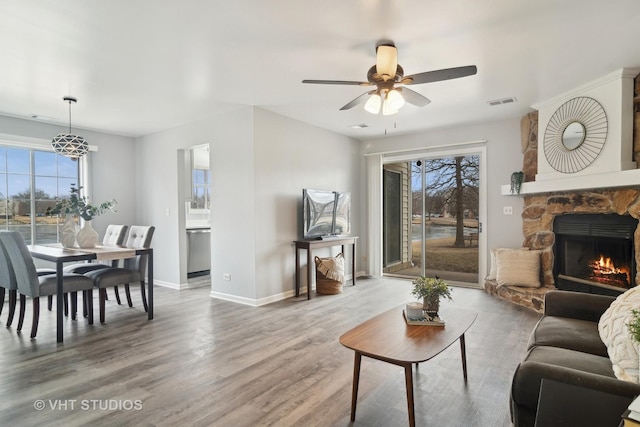 living room with ceiling fan, a stone fireplace, wood-type flooring, and a healthy amount of sunlight