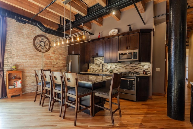 kitchen with brick wall, a towering ceiling, appliances with stainless steel finishes, light stone counters, and light hardwood / wood-style flooring