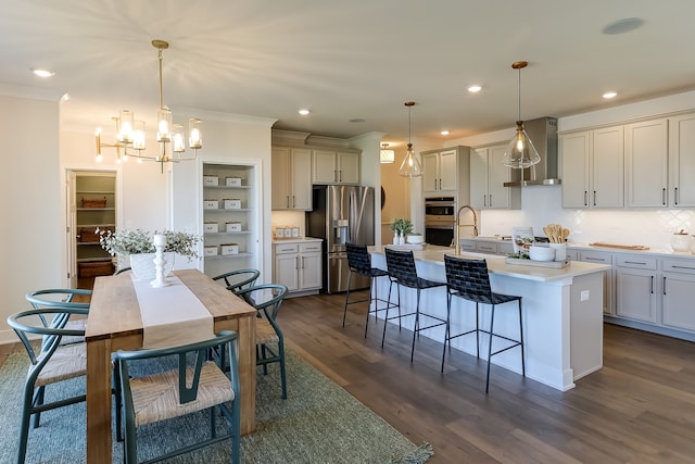 kitchen with pendant lighting, wall chimney range hood, stainless steel appliances, dark hardwood / wood-style floors, and a center island with sink