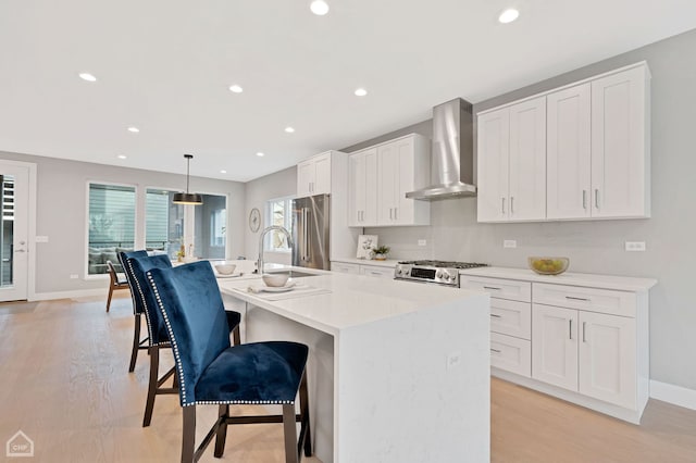 kitchen featuring wall chimney range hood, a kitchen island with sink, white cabinetry, hanging light fixtures, and stainless steel appliances