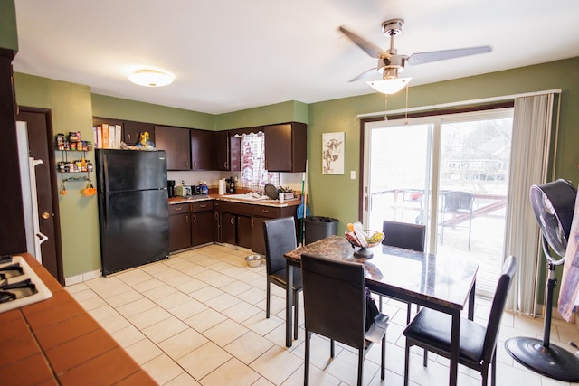 kitchen featuring light tile patterned floors, ceiling fan, black refrigerator, dark brown cabinetry, and white gas stovetop