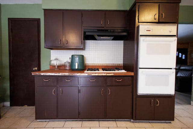 kitchen featuring exhaust hood, dark brown cabinets, light tile patterned floors, white appliances, and backsplash