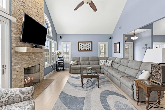 living room with ceiling fan, a stone fireplace, high vaulted ceiling, and light wood-type flooring