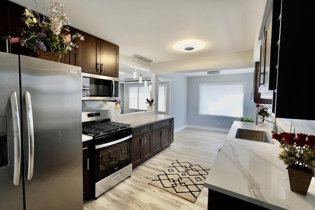 kitchen with sink, appliances with stainless steel finishes, dark brown cabinets, light stone counters, and light wood-type flooring