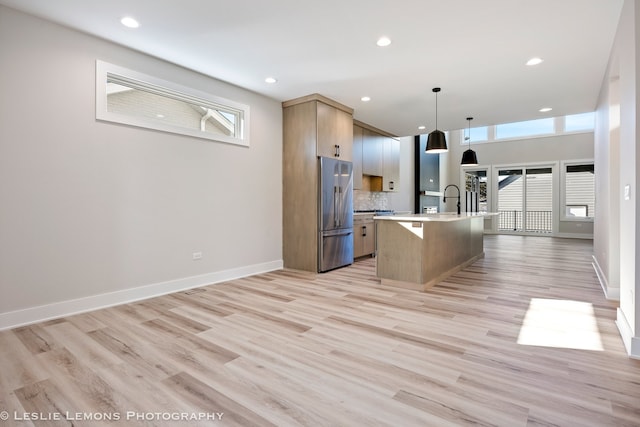 kitchen with light hardwood / wood-style flooring, hanging light fixtures, high quality fridge, an island with sink, and decorative backsplash