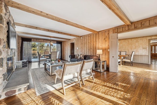 living room with a stone fireplace, wood walls, a chandelier, beam ceiling, and hardwood / wood-style floors