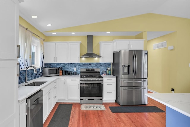 kitchen with vaulted ceiling, appliances with stainless steel finishes, sink, white cabinets, and wall chimney range hood