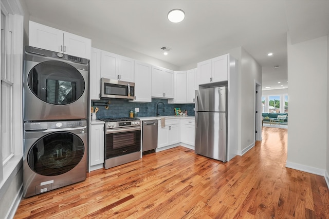 kitchen featuring light wood-type flooring, stainless steel appliances, stacked washing maching and dryer, decorative backsplash, and white cabinets