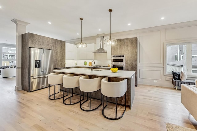 kitchen with decorative light fixtures, a center island with sink, appliances with stainless steel finishes, light hardwood / wood-style floors, and wall chimney range hood