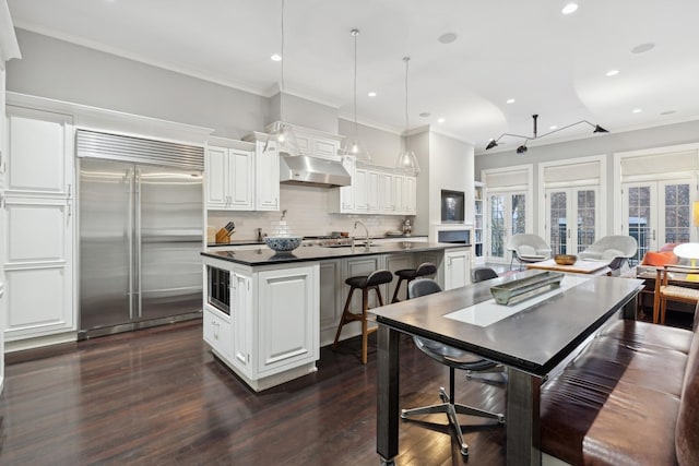 kitchen with a kitchen island with sink, built in appliances, white cabinets, and decorative light fixtures