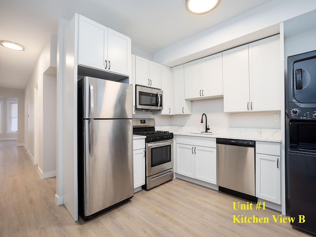 kitchen with stacked washing maching and dryer, appliances with stainless steel finishes, white cabinetry, sink, and light wood-type flooring