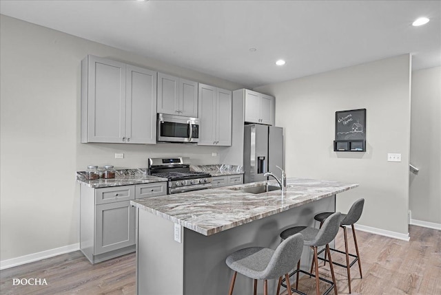 kitchen with sink, gray cabinetry, stainless steel appliances, light stone countertops, and a center island with sink