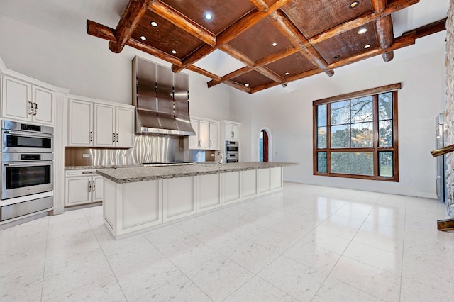 kitchen featuring coffered ceiling, white cabinetry, an island with sink, a towering ceiling, and light stone countertops