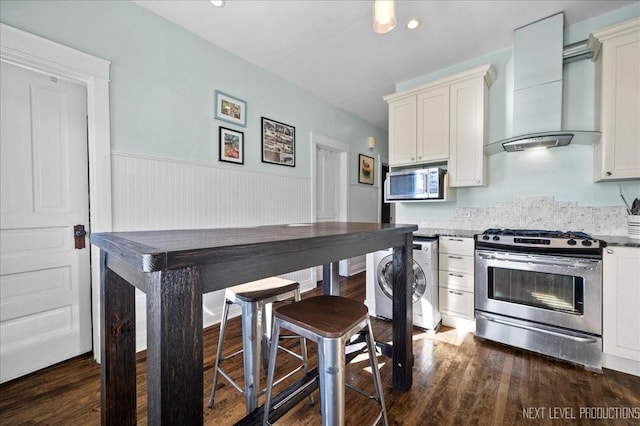 kitchen with washer / clothes dryer, wall chimney range hood, dark wood-type flooring, and stainless steel appliances