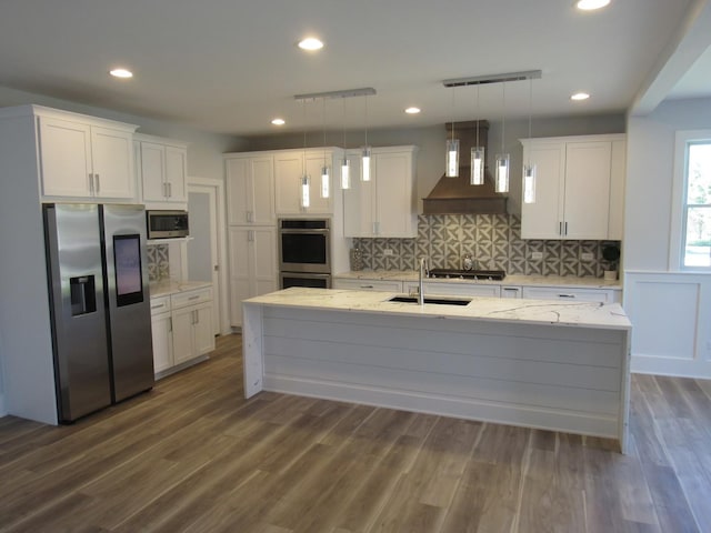 kitchen featuring white cabinetry, decorative light fixtures, a center island with sink, custom range hood, and stainless steel appliances