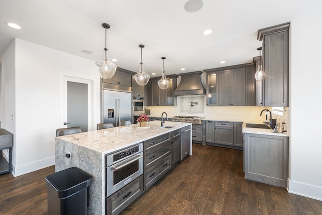 kitchen with appliances with stainless steel finishes, sink, custom exhaust hood, hanging light fixtures, and a kitchen island with sink