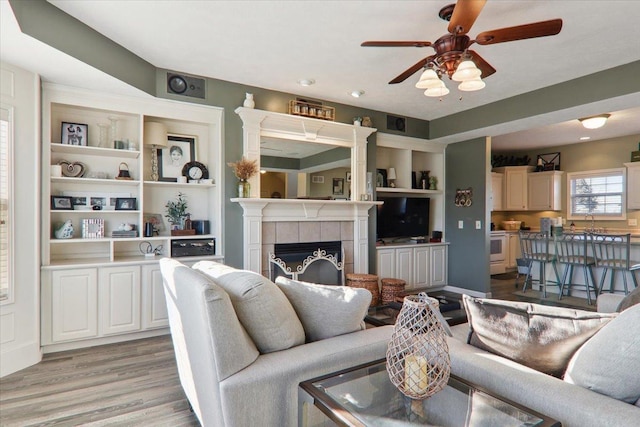 living room with a tiled fireplace, light hardwood / wood-style flooring, built in features, and ceiling fan