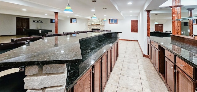 kitchen with a spacious island, a stone fireplace, hanging light fixtures, light tile patterned floors, and dark stone counters