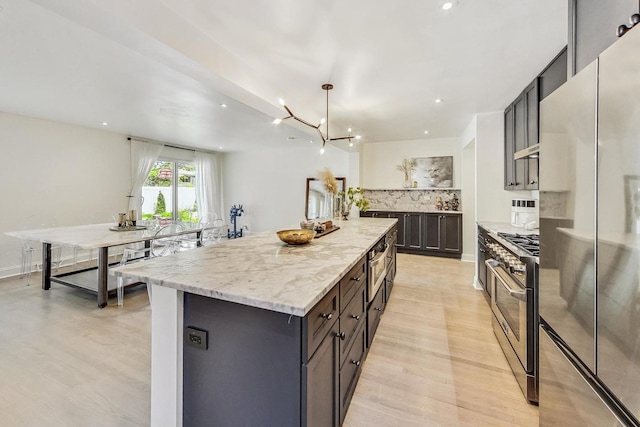 kitchen with backsplash, a chandelier, light stone counters, stainless steel appliances, and a center island with sink