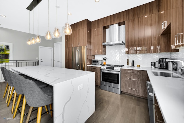 kitchen with backsplash, a kitchen bar, hanging light fixtures, stainless steel appliances, and wall chimney exhaust hood