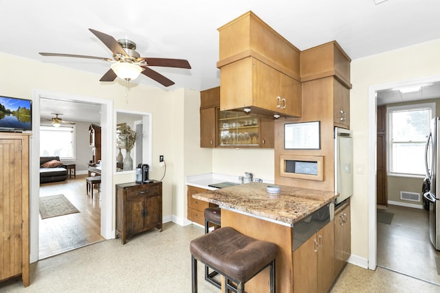 kitchen with a breakfast bar area, light stone counters, stainless steel refrigerator, kitchen peninsula, and ceiling fan