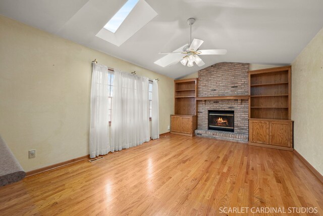 unfurnished living room with built in features, ceiling fan, vaulted ceiling with skylight, a fireplace, and light hardwood / wood-style floors