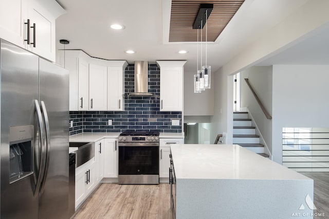 kitchen with wall chimney range hood, stainless steel appliances, hanging light fixtures, and white cabinets