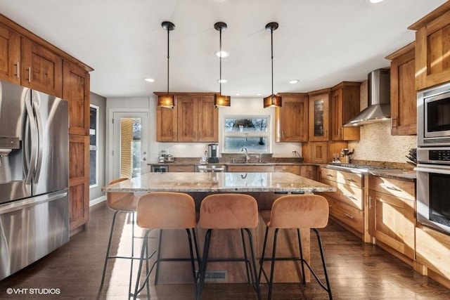 kitchen with wall chimney exhaust hood, hanging light fixtures, appliances with stainless steel finishes, a kitchen island, and light stone countertops