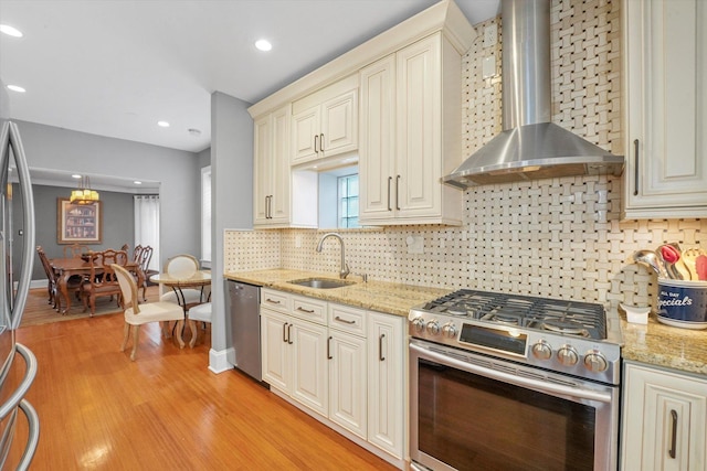 kitchen with sink, decorative light fixtures, appliances with stainless steel finishes, cream cabinets, and wall chimney range hood