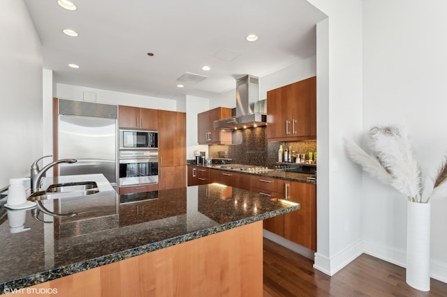 kitchen with dark stone counters, built in appliances, and wall chimney range hood