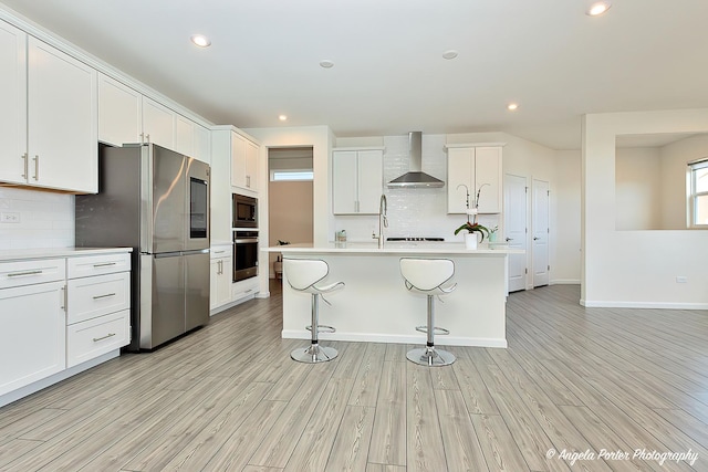 kitchen with white cabinets, wall chimney range hood, stainless steel appliances, a center island with sink, and light hardwood / wood-style flooring