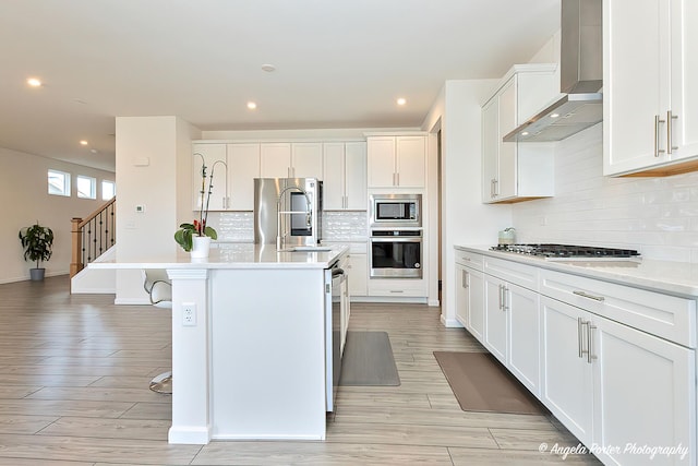 kitchen with wall chimney exhaust hood, a breakfast bar, white cabinetry, appliances with stainless steel finishes, and an island with sink