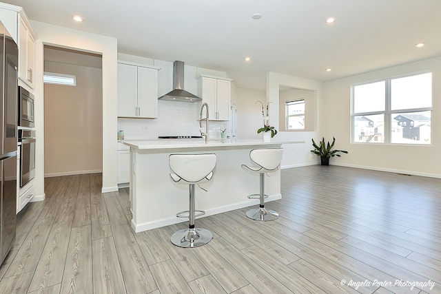 kitchen with white cabinetry, a center island with sink, appliances with stainless steel finishes, decorative backsplash, and wall chimney range hood