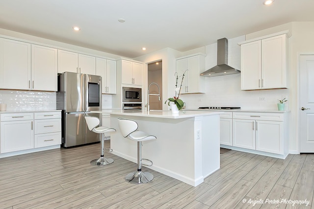 kitchen with white cabinetry, light hardwood / wood-style flooring, a center island with sink, appliances with stainless steel finishes, and wall chimney range hood