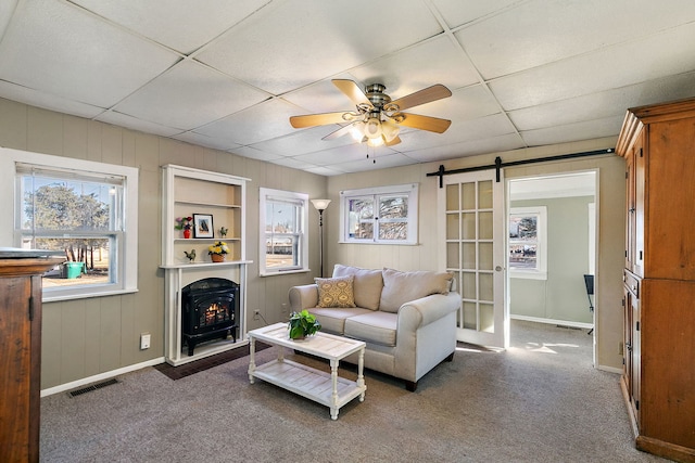 carpeted living room featuring a barn door, wooden walls, a drop ceiling, and ceiling fan