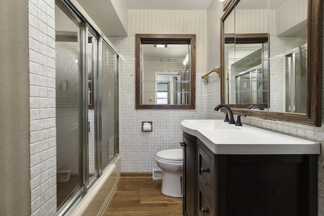 full bathroom with bath / shower combo with glass door, wood-type flooring, tile walls, vanity, and toilet