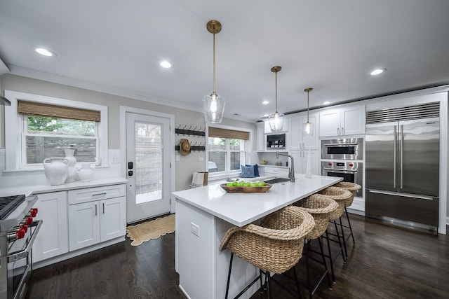 kitchen with sink, white cabinetry, hanging light fixtures, built in appliances, and a center island with sink