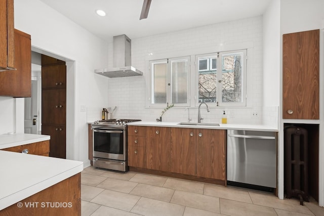 kitchen with brown cabinets, stainless steel appliances, light countertops, wall chimney range hood, and a sink