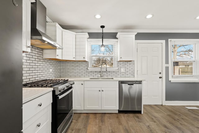 kitchen with sink, white cabinets, hanging light fixtures, stainless steel appliances, and wall chimney range hood