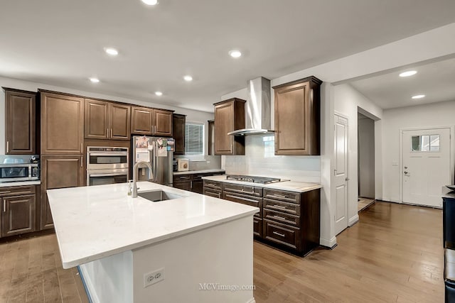 kitchen featuring sink, appliances with stainless steel finishes, a center island with sink, wall chimney exhaust hood, and light wood-type flooring