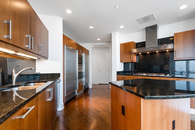 kitchen featuring a kitchen island, sink, dark stone counters, stainless steel appliances, and wall chimney exhaust hood